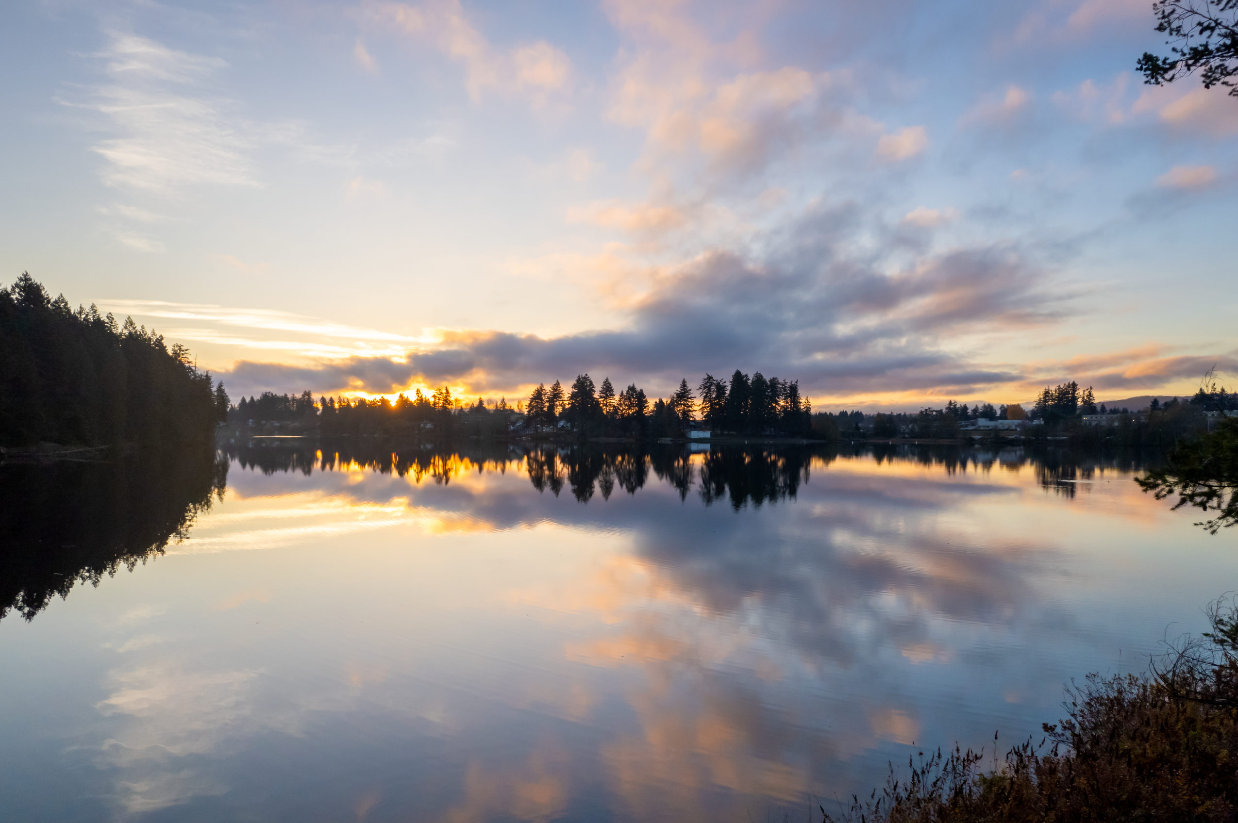 A tranquil sunset scene with soft clouds reflecting on a calm lake surrounded by tall evergreen trees.